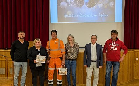 Sechs Personen stehen nebeneinander auf einer Bühne vor rotem Vorhang und Leinwand mit Jubelbild und Text. Es handelt sich um den Personalrat der Stadt Monheim und geehrte Jubilare der Stadt Monheim