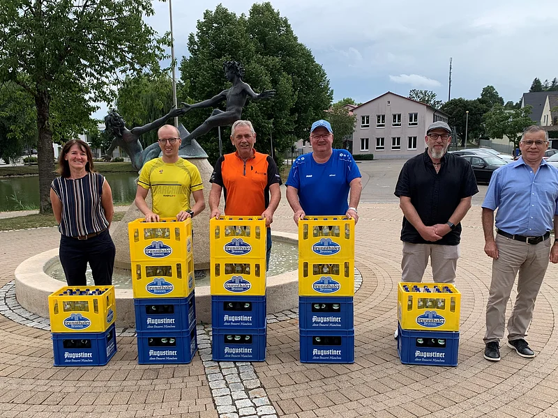 Sechs Personen stehen hinter Bierkästen vor einem Brunnen mit Skulptur auf einem gepflasterten Platz. Im Hintergrund sind Bäume, geparkte Fahrzeuge und ein kleiner Weiher.