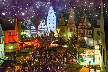Abendaufnahme des belebten Monheimer Weihnachtsmarktes in der Altstadt von Monheim: mit Fachwerkhäusern, Menschenmengen, Lichtern, Verkaufsständen und einer Bühne.