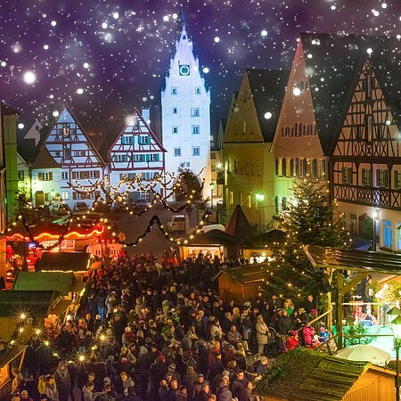 Abendaufnahme des belebten Monheimer Weihnachtsmarktes in der Altstadt von Monheim: mit Fachwerkhäusern, Menschenmengen, Lichtern, Verkaufsständen und einer Bühne.