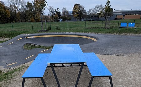 Blauer Picknicktisch mit Bänken auf Kiesfläche, davor verstreute rote Feuerwerkskörperreste. Verschmutzung an der Pumptrack-Anlage in Monheim.