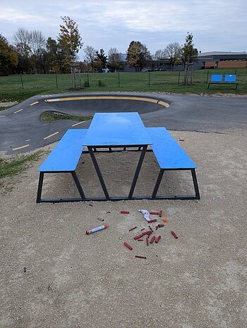 Blauer Picknicktisch mit Bänken auf Kiesfläche, davor verstreute rote Feuerwerkskörperreste. Verschmutzung an der Pumptrack-Anlage in Monheim.