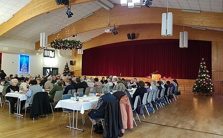 Menschen sitzen in mehreren Reihen an weißgedeckten Tischen in einem Saal mit einer Holzbalkendecke und Weihnachtsdekoration. Eine Bühne mit einem roten Vorhang ist im Hintergrund.