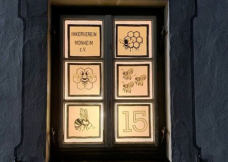 Beleuchtetes Adventskalenderfenster in sechs Bereiche unterteilt, darauf Bienenmotive, Waben und der Text „Imkerverein Monheim e.V.“ sowie die Zahl 15 rechts unten im Bild.