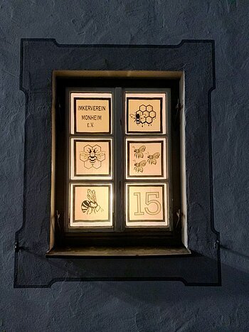 Beleuchtetes Adventskalenderfenster in sechs Bereiche unterteilt, darauf Bienenmotive, Waben und der Text „Imkerverein Monheim e.V.“ sowie die Zahl 15 rechts unten im Bild.