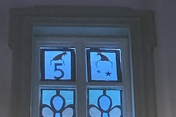 Adventskalenderfenster weihnachtlich dekoriert und von hinten beleuchtet mit zwei Engeldarstellungen, eine mit gelbem Stern, eine mit rotem Herz in der Körpermitte. Links oben im Eck Zahl 5 und rechts davon sind Sterne, beides mit einer Weihnachtsmütze darüber.