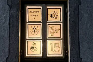 Beleuchtetes Adventskalenderfenster in sechs Bereiche unterteilt, darauf Bienenmotive, Waben und der Text „Imkerverein Monheim e.V.“ sowie die Zahl 15 rechts unten im Bild.