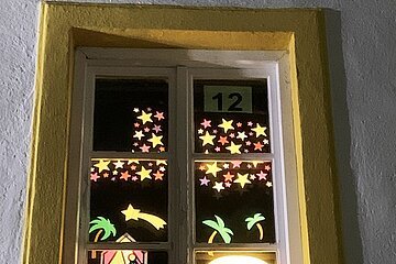 Adventskalenderfenster mit gelbem Rahmen zeigt beleuchtete Weihnachtsmotive mit Sternen, Palmen, Krippe und drei Königen. Oben rechts im Fenster die Zahl 12 auf Papier.
