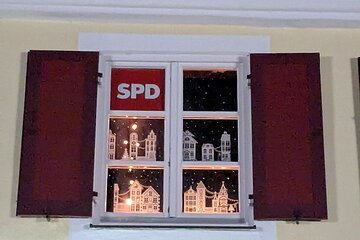 Adventskalenderfenster mit weißer Umrandung und geöffneten roten Fensterläden weihnachtlich gestaltet.  Beleuchtete Papierhäusern als Dekoration auf schwarzem Hintergrund mit Sternenhimmel. Oben links im Eck das Logo der SPD in rot-weiß.
