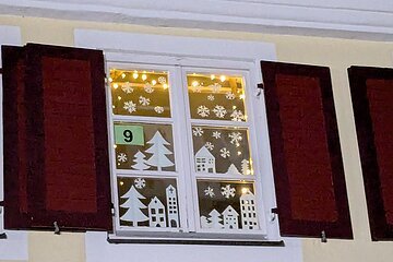 Adventskalenderfenster mit Weihnachtsdekoration aus Papier in Form von Häusern, Tannenbäumen und  Schneeflocken. Oben eine Lichterkette und in der Mitte des Fensters ein Schild mit der Zahl 9.