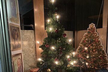 Adventskalenderfenster weihnachtlich dekoriert mit einem beleuchtetem Baum und roten Christbaumkugeln drauf. Zwei Spielzeug-Feuerwehrautos, eingepackten Geschenken und drei Portraits. Rechts im Bild ist ein geschmückter Tannenbaum aus Pappe aufgestellt. Auf der Fensterscheibe ist ein Schild aus Papier mit der Zahl 8 angebracht.