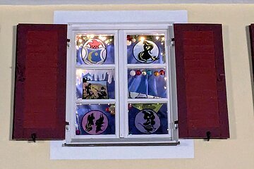 Adventskalenderfenster mit weißen Rahmen und roten Fensterläden weihnachtlich dekoriert. Bunte von einer Lichterkette beleuchtete Bilder mit blauem Vorhang dahinter, Christbaumkugeln und dekorativer Schnee hängen herunter. Links oben im Fenster das Logo des Faschingsvereins Gailachia Monheim.