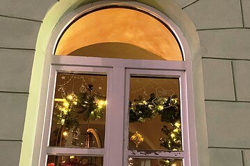 Adventskalenderfenster weihnachtlich dekoriert mit Weihnachtsbeleuchtung, einem Holzschlitten, Girlanden, Schneeflocken, einer Kerze, einer Figur eines Schützen und einer Siegesscheibe eines Schützenvereins.