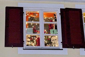 Adventskalender mit weißem Rahmen und geöffneten roten Fensterläden weihnachtlich dekoriert. Innen ist Weihnachtsbeleuchtung und Dekoration wie rote Christbaumkugeln, Sterne oben und rote Geschenkschleifen. In der Mitte des Fensters rechts ist die Zahl 1 auf Papier angebracht. Darunter ein Portrait eines Mannes zu sehen.