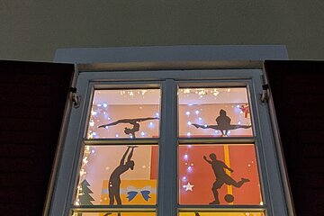 Adventskalenderfenster mit weißem Rahmen und roten Fensterläden weihnachtlich dekoriert. Darin fünf unterschiedliche Silhouetten von Sportlern beim Sport und links unten im Eck der Text „Frohe Weihnachten und ein gutes neues Jahr“ mit dem TSV Monheim Logo rechts daneben. Um das komplette Fenster eine leuchtende Lichterkette.