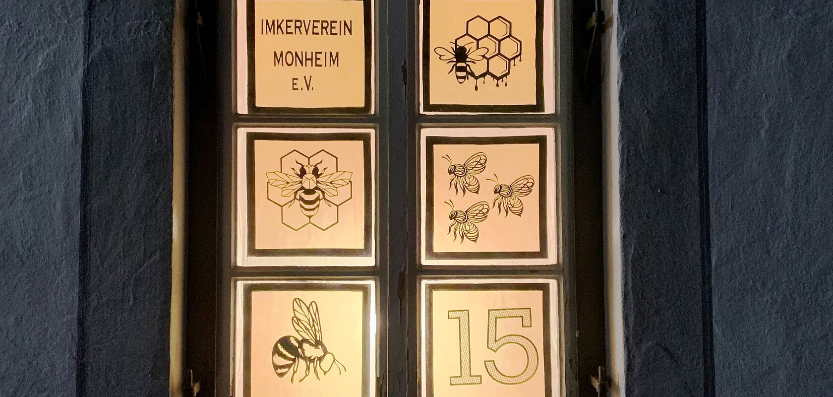 Beleuchtetes Adventskalenderfenster in sechs Bereiche unterteilt, darauf Bienenmotive, Waben und der Text „Imkerverein Monheim e.V.“ sowie die Zahl 15 rechts unten im Bild.