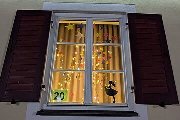 Adventskalenderfenster mit weißer Umrandung und geöffneten roten Fensterläden weihnachtlich dekoriert. Innen bunte Sterne umgeben von einer Lichterkette und unten rechts im Eck in schwarz die Silhouette eines Mädchens mit Kleid. Unten links im Eck die Zahl 20 auf Papier mit grünem Hintergrund.
