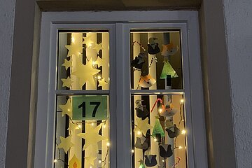 Adventskalenderfenster mit einer Umrandung in beige weihnachtlich dekoriert. Darin von oben hängend gebastelte Weihnachtsdeko wie Papiersterne, Kerzen, Tannenbäume und Tiergesichter. In der Mitte des Fensters die Zahl 17 auf grünem Hintergrund. Das Fenster ist mit einer Lichterkette beleuchtet.