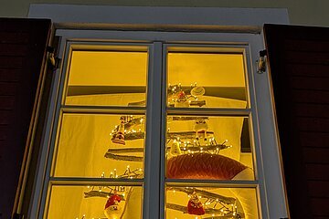 Adventskalenderfenster mit weißem Rahmen und geöffneten roten Fensterläden weihnachtlich dekoriert. Darin Weihnachtsdeko wie  ein Tannenbaum aus Holz mit einer Lichterkette, der mit Figuren wie Weihnachtsmänner, Leuchtturm und Engeln dekoriert ist. Rechts unten auf dem Fensterbrett steht ein Lebkuchenmann. Hinter dem Lebkuchenmann ist ein rot-weißer Rettungsring innen sichtbar.