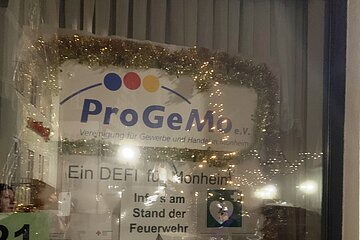 Adventskalenderfenster mit Lametta weihnachtlich dekoriert mit einer Darstellung zur geplanten Aktion der ProGeMo Monheim. Auf dem Plakat oben das Logo der ProGeMo-Monheim, darunter der Schriftzug "Ein DEFI für Monheim", darunter ein Defibrillator und um den Defibrillator sind Informationsplakate und Symbole angebracht. Auf der Scheibe links ist ein Blatt mit der Zahl 21 auf Papier angebracht. Auf dem Fensterbrett außen ist ein bepflanzter Blumentopf.