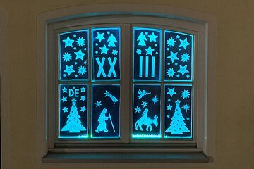 Adventskalenderfenster mit leuchtenden Weihnachtsmotiven wie Sternen, Engeln, Tannenbäumen, einer Krippenszene,  römischen Zahlen und dem Schriftzug „DE LUMINA“ in blauem Licht.