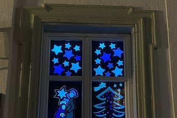 Adventskalenderfenster weihnachtlich dekoriert auf schwarzem Hintergrund mit leuchtenden Sternen oben im Fenster, einem Weihnachtsbaum rechts im Fenster, Krippenfiguren links davon und dem Schriftzug "Die Monheimer Ministranten wünschen gesegnete Weihnachten." Unter dem Tannenbaum die Zahl 24. Alles in hell- und dunkelblau und von hinten beleuchtet.