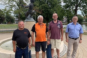 Vier Männer stehen vor einem Brunnen mit einer bronzenen Skulptur auf einem gepflastertem Platz. Im Hintergrund sind Bäume und ein kleiner Weiher.