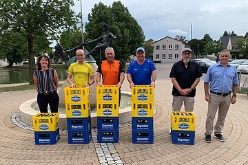 Sechs Personen stehen hinter Bierkästen vor einem Brunnen mit Skulptur auf einem gepflasterten Platz. Im Hintergrund sind Bäume, geparkte Fahrzeuge und ein kleiner Weiher.