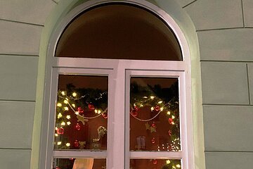 Adventskalenderfenster mit Weihnachtsbeleuchtung, von oben hängenden roten Kugeln, Lebkuchenfiguren, Musikinstrumenten und einem Plüschelefanten mit einer weißen Mütze im Inneren.