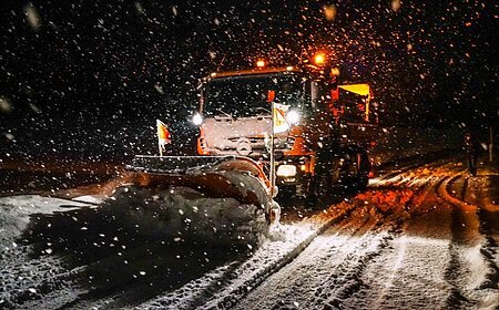 Ein Schneepflug räumt nachts eine verschneite Straße, während es schneit.