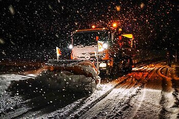 Ein Schneepflug räumt nachts eine verschneite Straße, während es schneit.