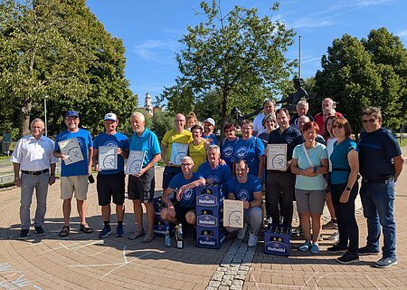 Gruppenfoto von Personen, die im Freien auf einem gepflastertem Platz stehen, einige halten Urkunden in den Händen. Neben der Gruppe stehen vier blaue Getränkekisten. Im Hintergrund sind Bäume, blauer Himmel, ein Kirchenturm und eine Statue, bei Sonnenschein.