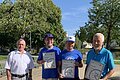 Vier Männer stehen im Freien auf einem gepflastertem Platz, drei halten Urkunden in den Händen, neben ihnen rechts im Bild stehen zwei blaue Getränkekisten. Im Hintergrund sind Bäume, blauer Himmel und ein Teich.