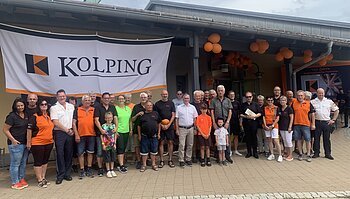 Gruppenfoto von etwa 30 Personen vor einem Gebäude auf einem gepflasterten Platz mit einem großen Kolping-Banner über der Gruppe und orangen Luftballons rechts im Bild.