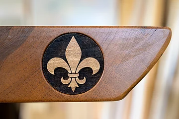 Das Logo des Möbelgeschäfts "Meßmer" in Monheim auf einem Stück Holz in braun. Das Logo enthält ein eingraviertes goldfarbenes Lilien-Symbol in einem schwarzen Kreis.