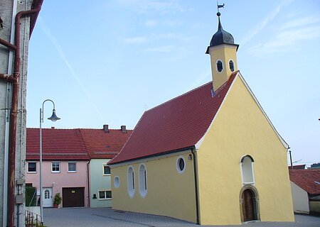 Gelbe Kapelle, die Peterskapelle in Monheim mit rotem Dach und einem Turm, umgeben von Wohnhäusern mit roten Dächern. Im Hintergrund blauer Himmel.