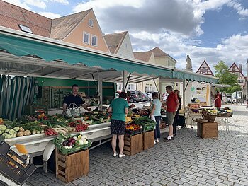 Marktstand des Wochenmarkts in Monheim mit Obst und Gemüse in der Auslage auf dem gepflastertem Marktplatz. Mehrere Personen kaufen dort ein. Fachwerkhäuser im Hintergrund und bewölkter Himmel.