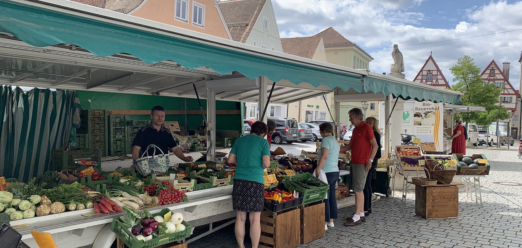 Marktstand des Wochenmarkts in Monheim mit Obst und Gemüse in der Auslage auf dem gepflastertem Marktplatz. Mehrere Personen kaufen dort ein. Fachwerkhäuser im Hintergrund und bewölkter Himmel.