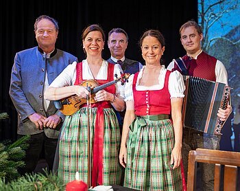 Fünf Personen in traditioneller Tracht, im Vordergrund zwei Frauen mit Geige und Dirndl, ein Mann mit Akkordeon rechts, vor dunklem Hintergrund.