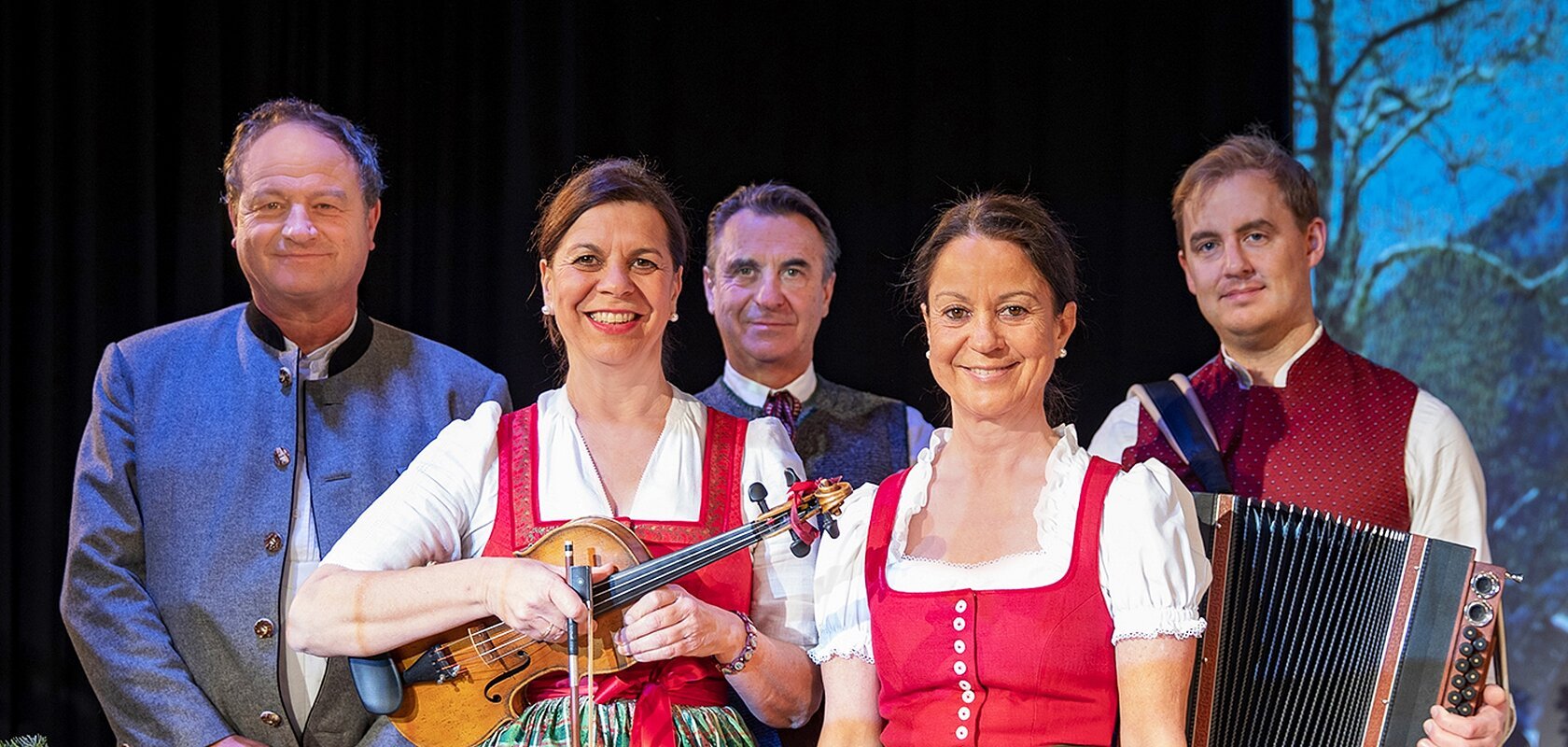 Fünf Personen in traditioneller Tracht, im Vordergrund zwei Frauen mit Geige und Dirndl, ein Mann mit Akkordeon rechts, vor dunklem Hintergrund.
