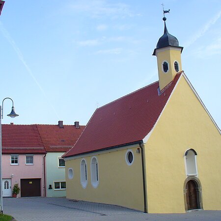 Gelbe Kapelle, die Peterskapelle in Monheim mit rotem Dach und einem Turm, umgeben von Wohnhäusern mit roten Dächern. Im Hintergrund blauer Himmel.