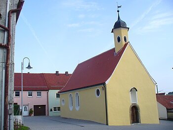 Gelbe Kapelle, die Peterskapelle in Monheim mit rotem Dach und einem Turm, umgeben von Wohnhäusern mit roten Dächern. Im Hintergrund blauer Himmel.