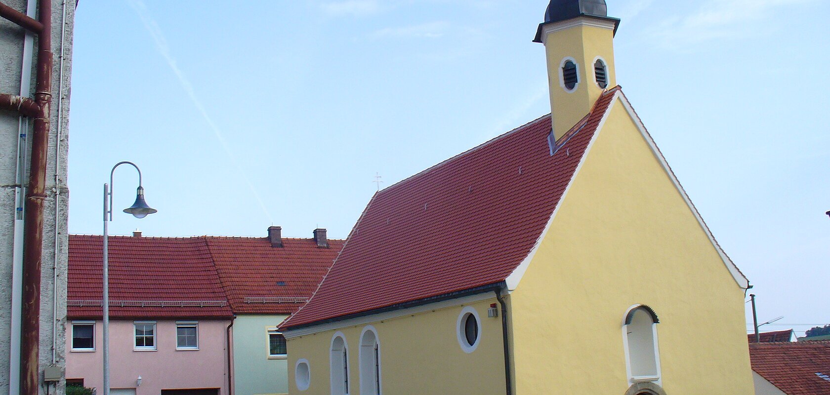 Gelbe Kapelle, die Peterskapelle in Monheim mit rotem Dach und einem Turm, umgeben von Wohnhäusern mit roten Dächern. Im Hintergrund blauer Himmel.