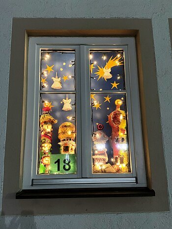Adventskalenderfenster mit einer Umrandung in beige weihnachtlich dekoriert. Darin mit Weihnachtsbeleuchtung beleuchtete Engelsfiguren, eine Sternenschnuppe und Sternen. Unten links die Zahl 18 auf grünem Hintergrund.