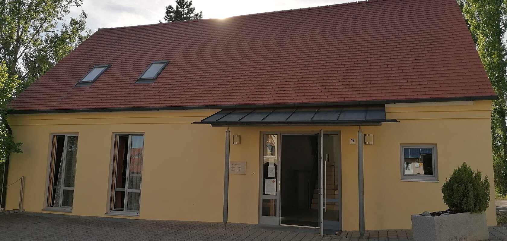 Das Haus Martin Luther in Monheim, ein evangelisches Gemeindehaus: Ein gelbes Gebäude mit rotem Satteldach, zwei Fenstern und einer offenen Eingangstür, umgeben von Bäumen. Vor dem Haus steht ein bepflanztes Beet.