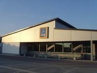 Gebäude mit schrägem Dach und ALDI-Süd Logo an der Fassade bei klarem Himmel. Vor den Fenstern des Gebäudes stehen viele zusammen geschobene Einkaufswägen aus Metall. Im Vordergrund ein Parkplatz.