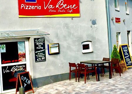 Außenansicht einer grau gestrichenen Pizzeria mit Tischen, Stühlen und mehreren Schildern mit Öffnungszeiten und Angeboten vor dem Gebäude. Zwei bepflanzte Töpfe stehen auf dem gepflastertem Platz.
