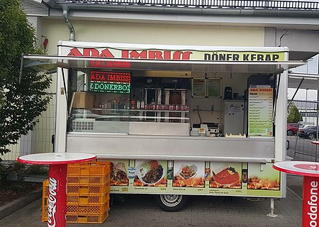 Imbisswagen mit geöffnetem Verkaufsfenster und zwei Stehtischen davor. Auf dem Wagen oben die Beschriftung „ADA Imbiss - Döner Kebab“ sichtbar unter dem geöffnetem Fenster verschiedene Abbildungen der erhältlichen Gerichte abgebildet. Links im Bild ein Baum, rechts ein Teil eines Parkplatzes zu sehen.