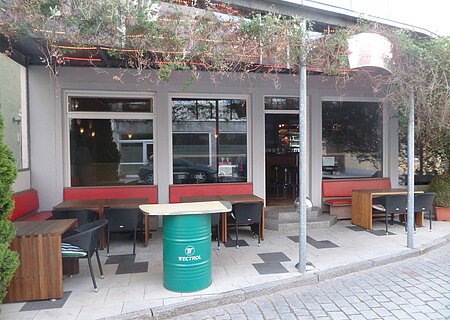 Außenbereich einer Kneipe mit roten Sitzbänken, Holztischen und schwarzen Stühlen. Vor dem Gebäude ein grünes Fass als Tisch auf einem gepflastertem Platz aufgestellt. Am Dach entlang verläuft ein grünes Gewächs. Die Überdachung ist mit rot-gelben LED-Streifen beleuchtet.