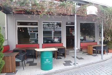 Außenbereich einer Kneipe mit roten Sitzbänken, Holztischen und schwarzen Stühlen. Vor dem Gebäude ein grünes Fass als Tisch auf einem gepflastertem Platz aufgestellt. Am Dach entlang verläuft ein grünes Gewächs. Die Überdachung ist mit rot-gelben LED-Streifen beleuchtet.